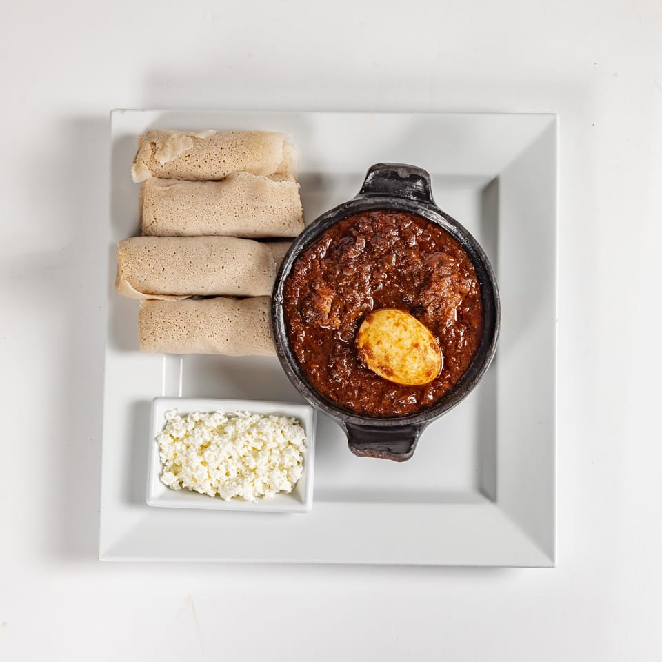 Overhead view of rolled injera, spicy stew topped with a boiled egg, and cottage cheese on a white plate.
