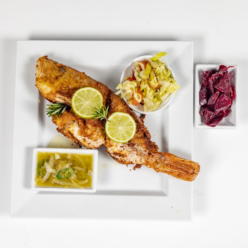 Golden fried whole fish garnished with lime and rosemary, served with salad, green sauce, and beet relish on a white plate.