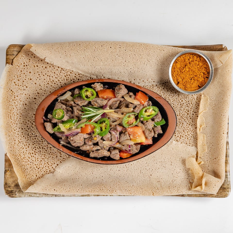 Ethiopian-style beef stew with peppers, tomatoes, and onions served on injera with a bowl of spices.