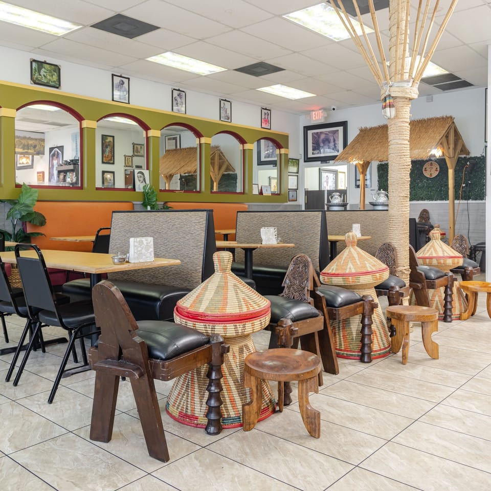 Spacious restaurant dining room with woven Ethiopian tables, wooden chairs, orange booths, arched decor, and thatched hut accents.