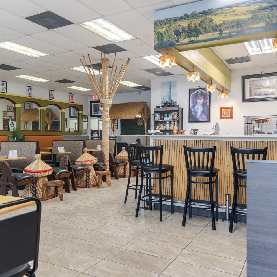 Spacious restaurant dining room with bamboo-paneled counter, woven stools, tropical decor, artwork, and empty tables.