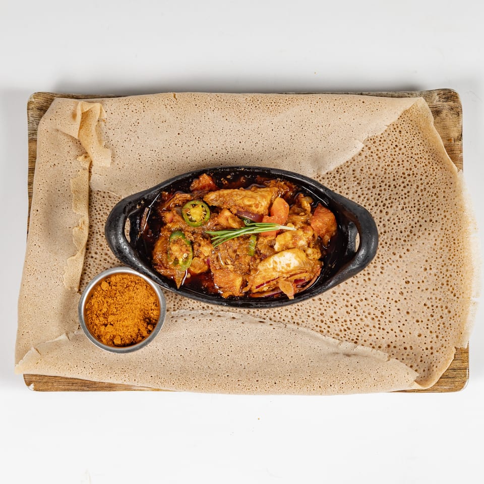 Spicy chicken stew garnished with jalapeños and rosemary in a black skillet, served on Ethiopian injera with dipping spice.