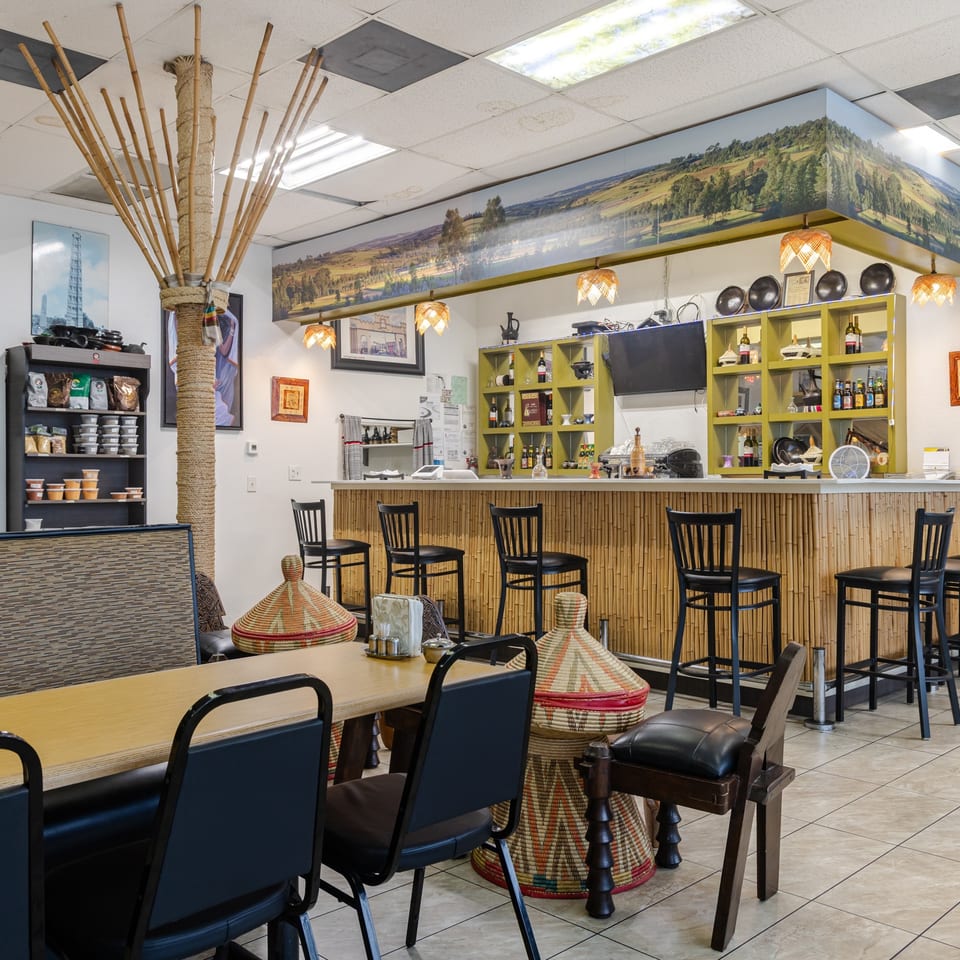 Bright restaurant interior with bamboo bar, colorful woven baskets, high-top seating, decorative shelves, and countryside mural.