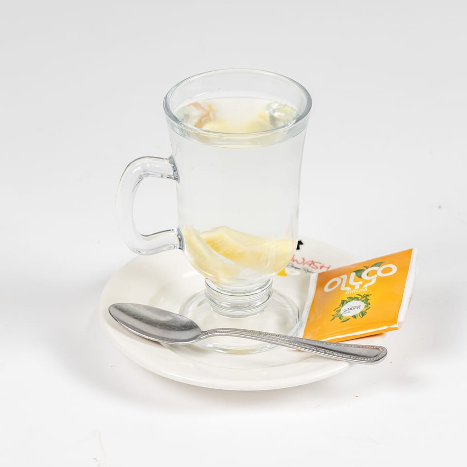 Glass mug of hot ginger tea with lemon slices, served on a saucer with a spoon and tea packet.