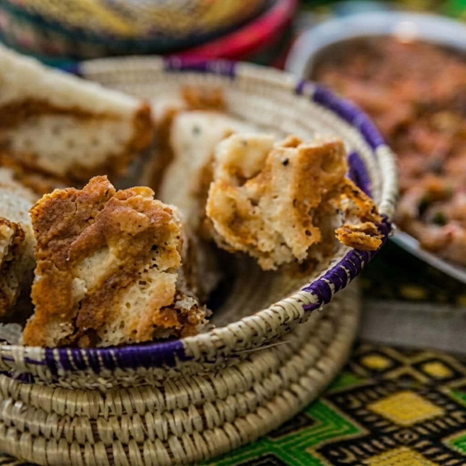 Chunks of crusty bread fill a woven basket beside a platter of saucy stew and tomato.