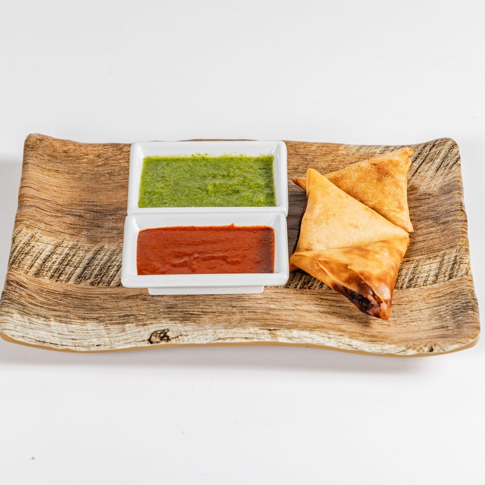 Three golden fried samosas served on a wooden platter with green and red dipping sauces.
