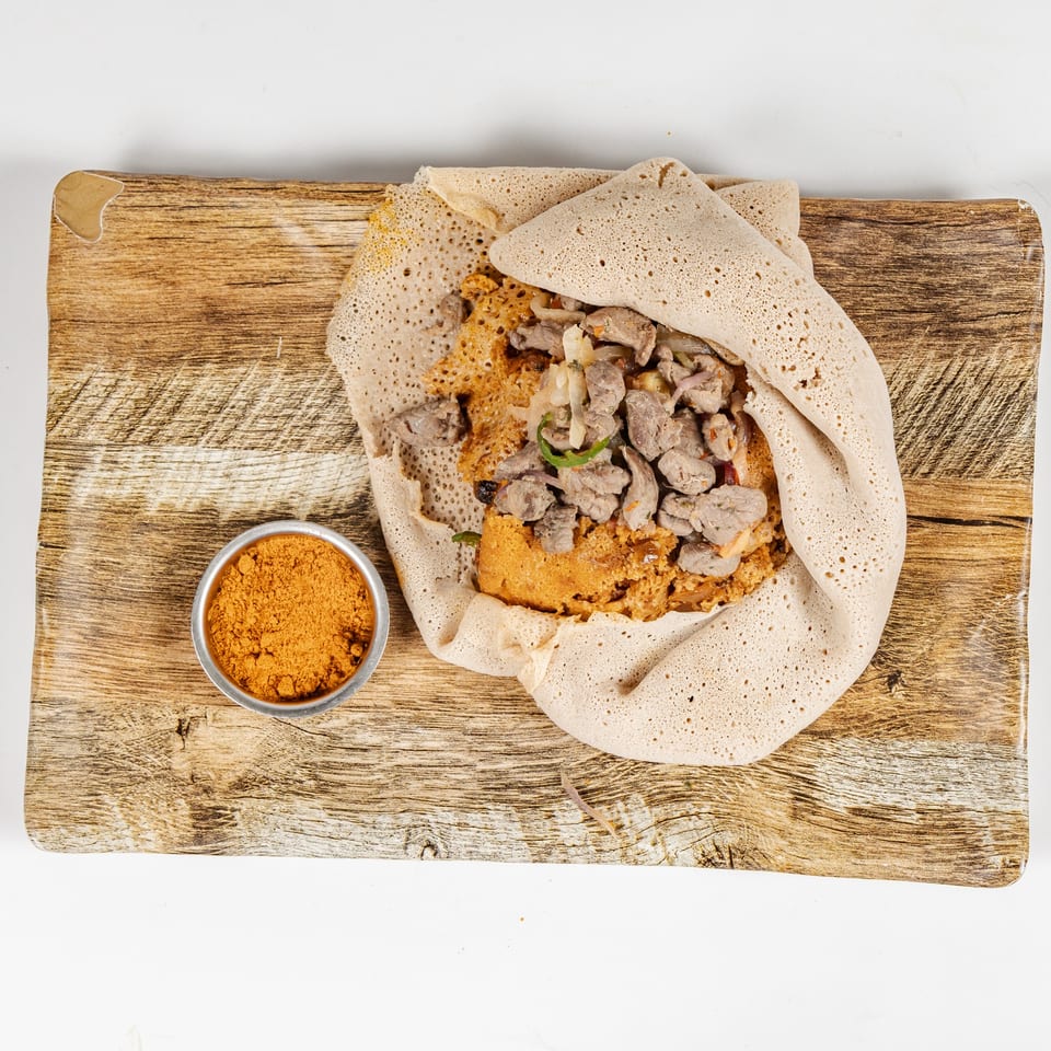 Ethiopian-style injera wraps surround seasoned meat and vegetables on a wooden serving board with a side of spice powder.