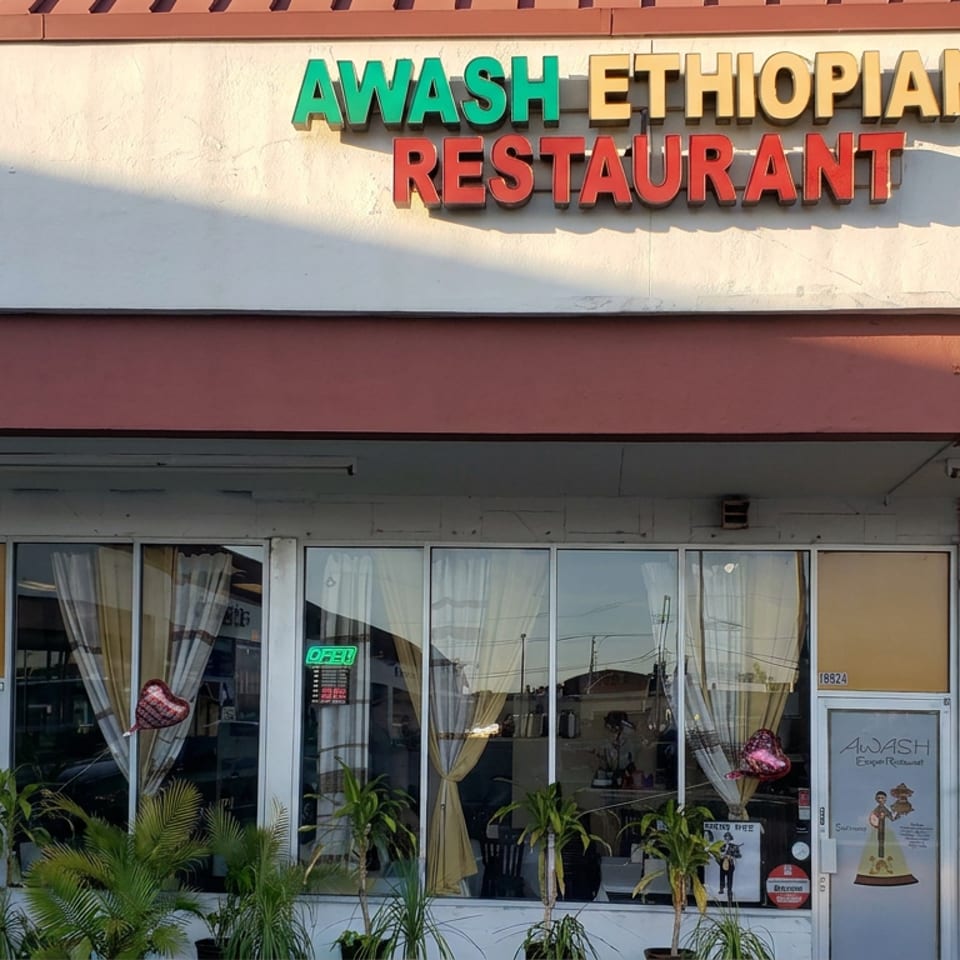 Exterior of Awash Ethiopian Restaurant featuring colorful signage, glass windows, curtains, potted palms, and a partially visible entrance.