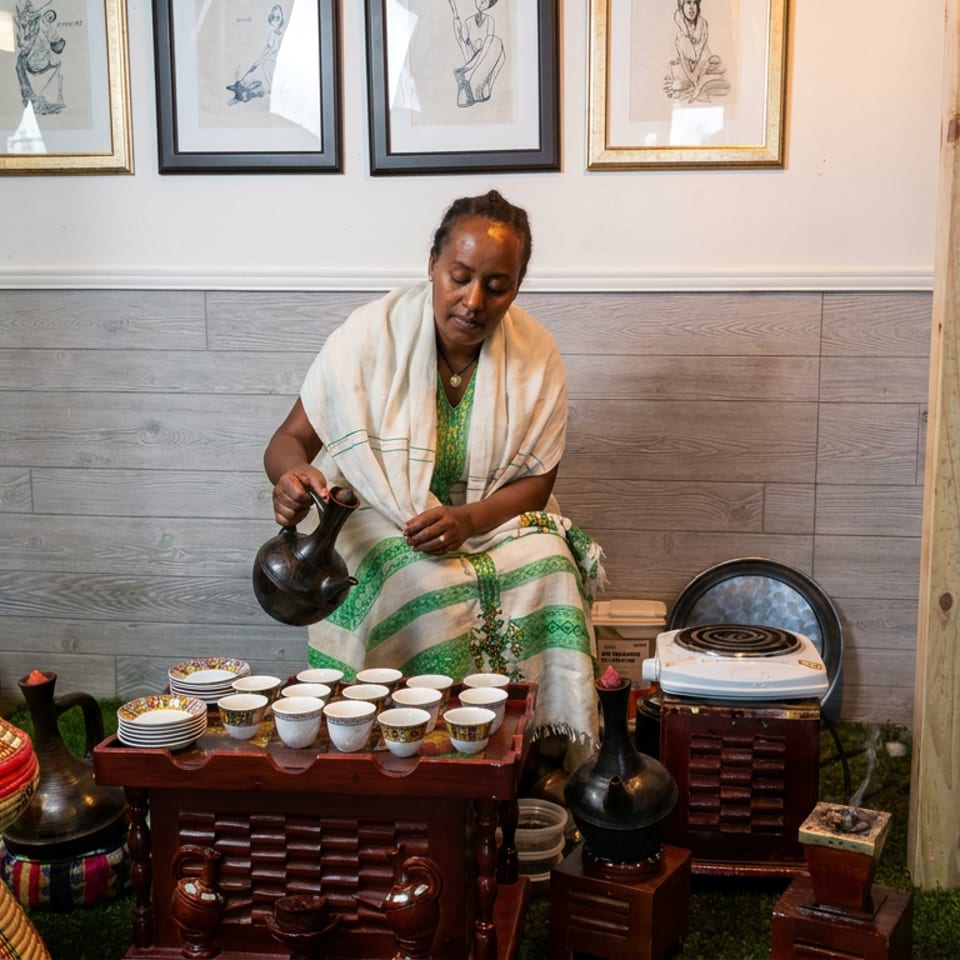 Woman in traditional attire pours coffee from a black pot beside a tray of cups in an Ethiopian coffee ceremony setting.