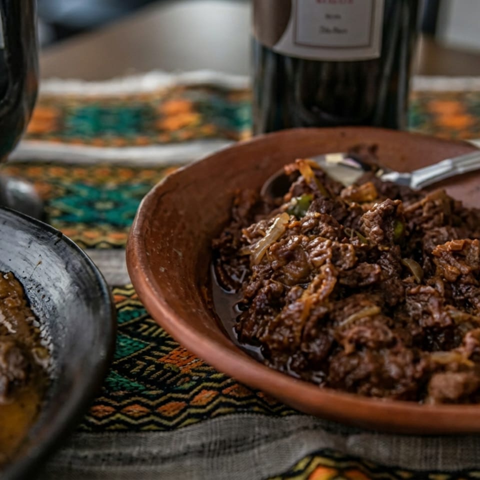 Rustic bowls of saucy meat dishes with vegetables, spoons, a wine bottle, and a patterned tablecloth.