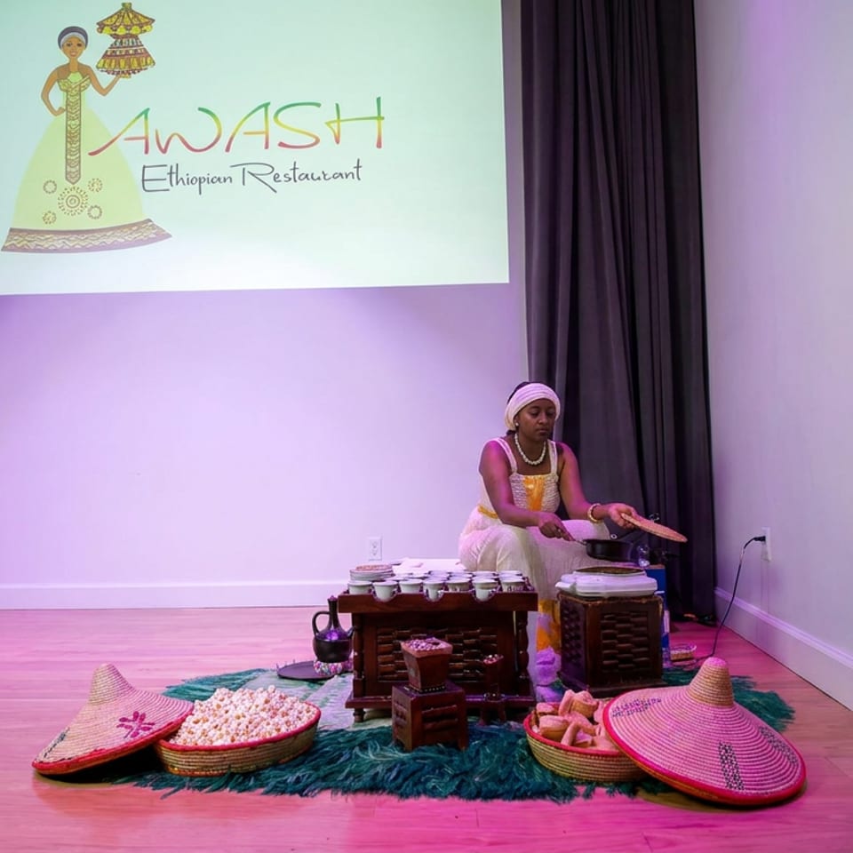 Woman in traditional Ethiopian attire performs a coffee ceremony beside woven baskets, popcorn, serving trays, and colorful hats.
