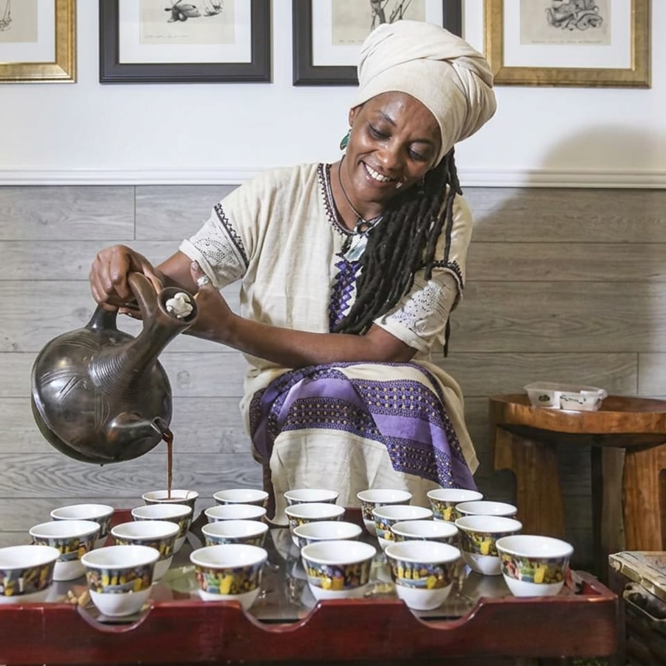 Smiling woman in traditional attire pours coffee from a black pot into cups arranged on a serving table.