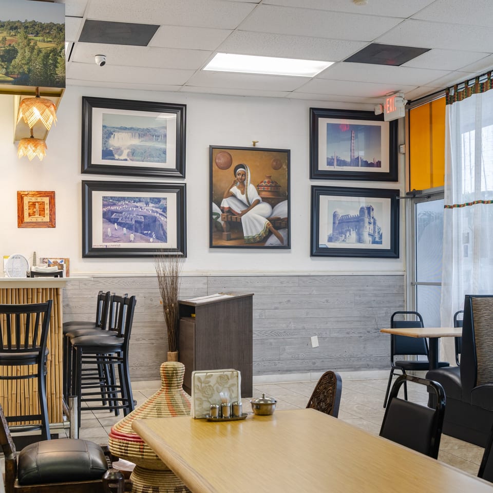 Bright restaurant dining room with black chairs, wooden tables, Ethiopian artwork, colorful woven decor, and a glass entrance.