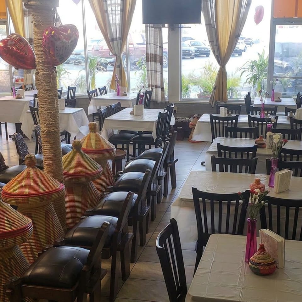 Bright restaurant dining room with white tablecloths, black chairs, woven decorative baskets, heart balloons, and large street-facing windows.