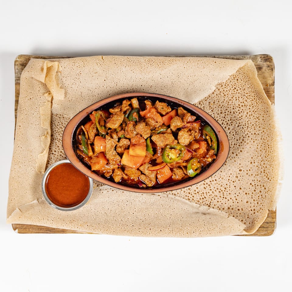 Spicy meat and vegetables served in a clay dish atop Ethiopian injera, with a small bowl of sauce.