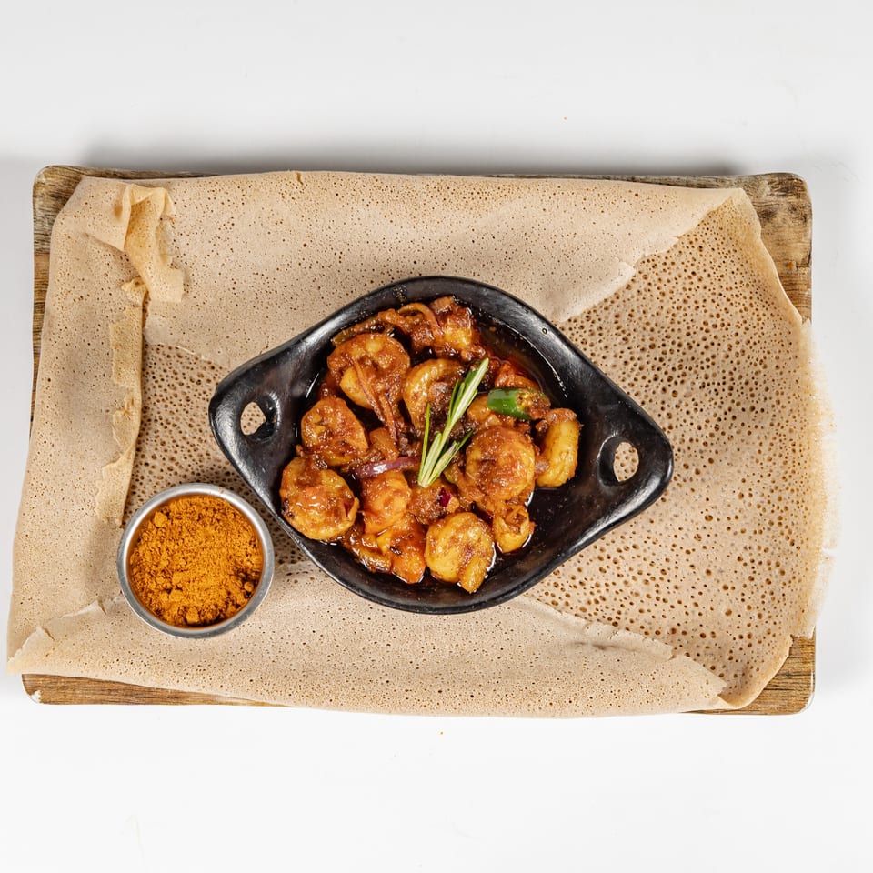 Ethiopian injera flatbread surrounds a black dish of spicy shrimp stew, garnished with herbs and served with berbere spice.