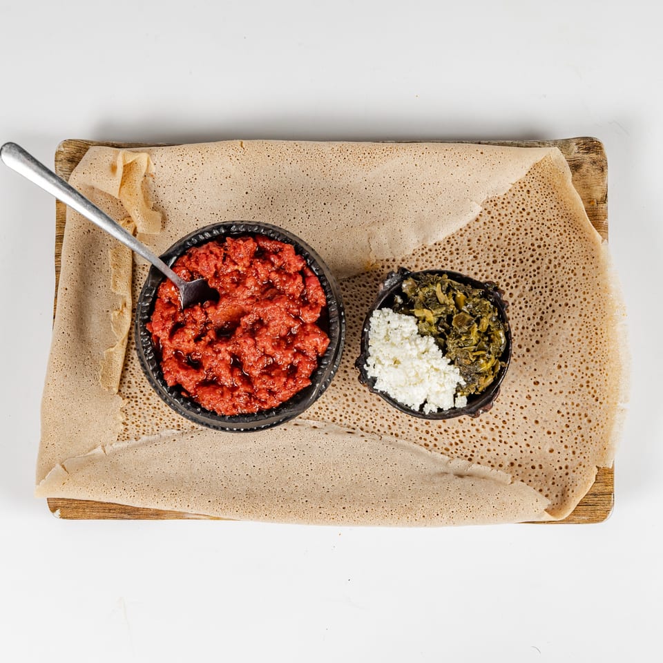 Overhead view of Ethiopian injera bread with bowls of spicy red stew, collard greens, and cottage cheese on a wooden platter.