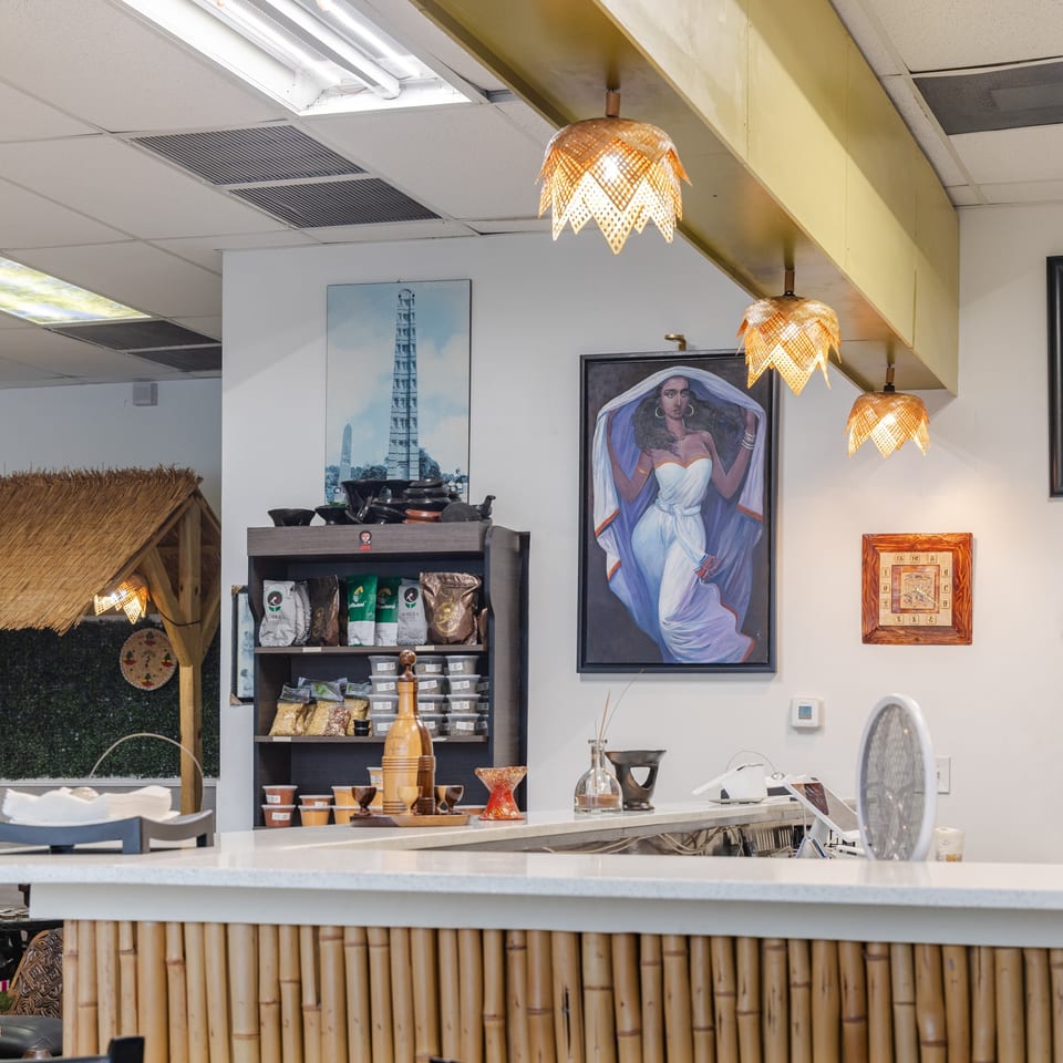 Restaurant interior with bamboo-fronted counter, woven pendant lights, cultural artwork, packaged goods, and seating area.