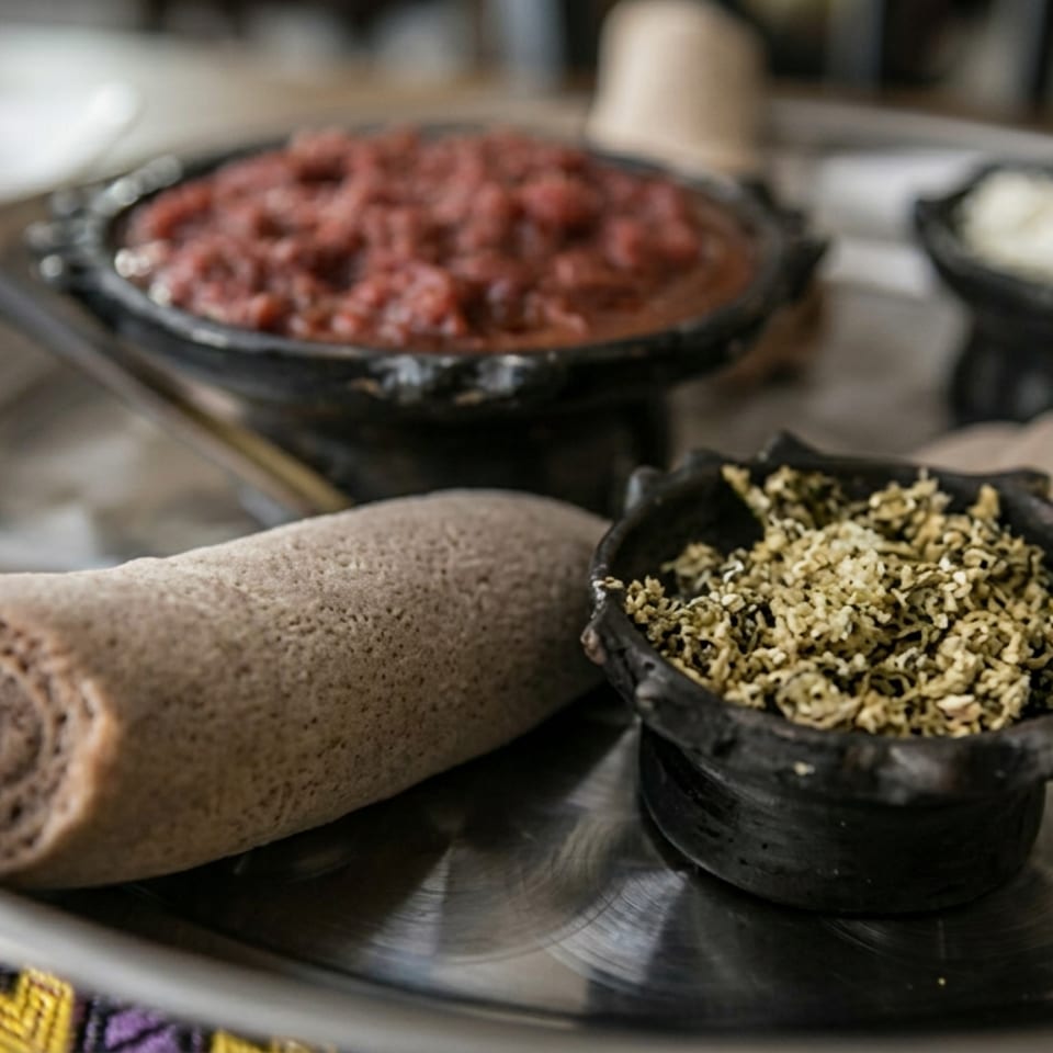 Ethiopian injera rolls surround black bowls of spiced vegetables, minced meat, and cottage cheese on a metal serving tray.