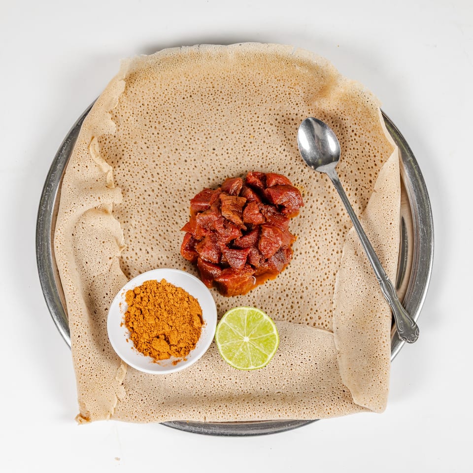 Ethiopian injera flatbread topped with spicy red meat, berbere spice, lime, and a spoon on a metal platter.