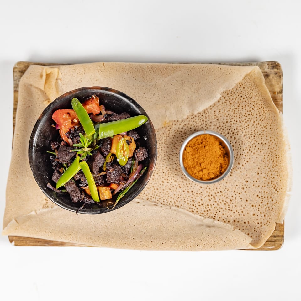 Overhead view of spiced beef with peppers and tomatoes in a black bowl, served on injera with dipping spice.